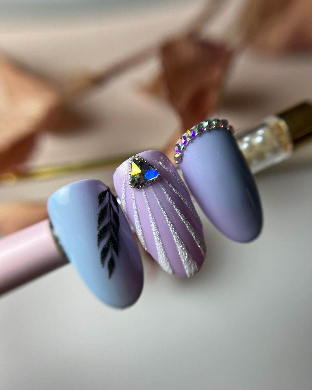 Trzy sztuczne paznokcie w odcieniach fioletu: jeden z czarnym wzorem liścia, drugi z srebrnymi paskami i trójkątnym kryształem, trzeci ozdobiony rzędem małych kryształków.