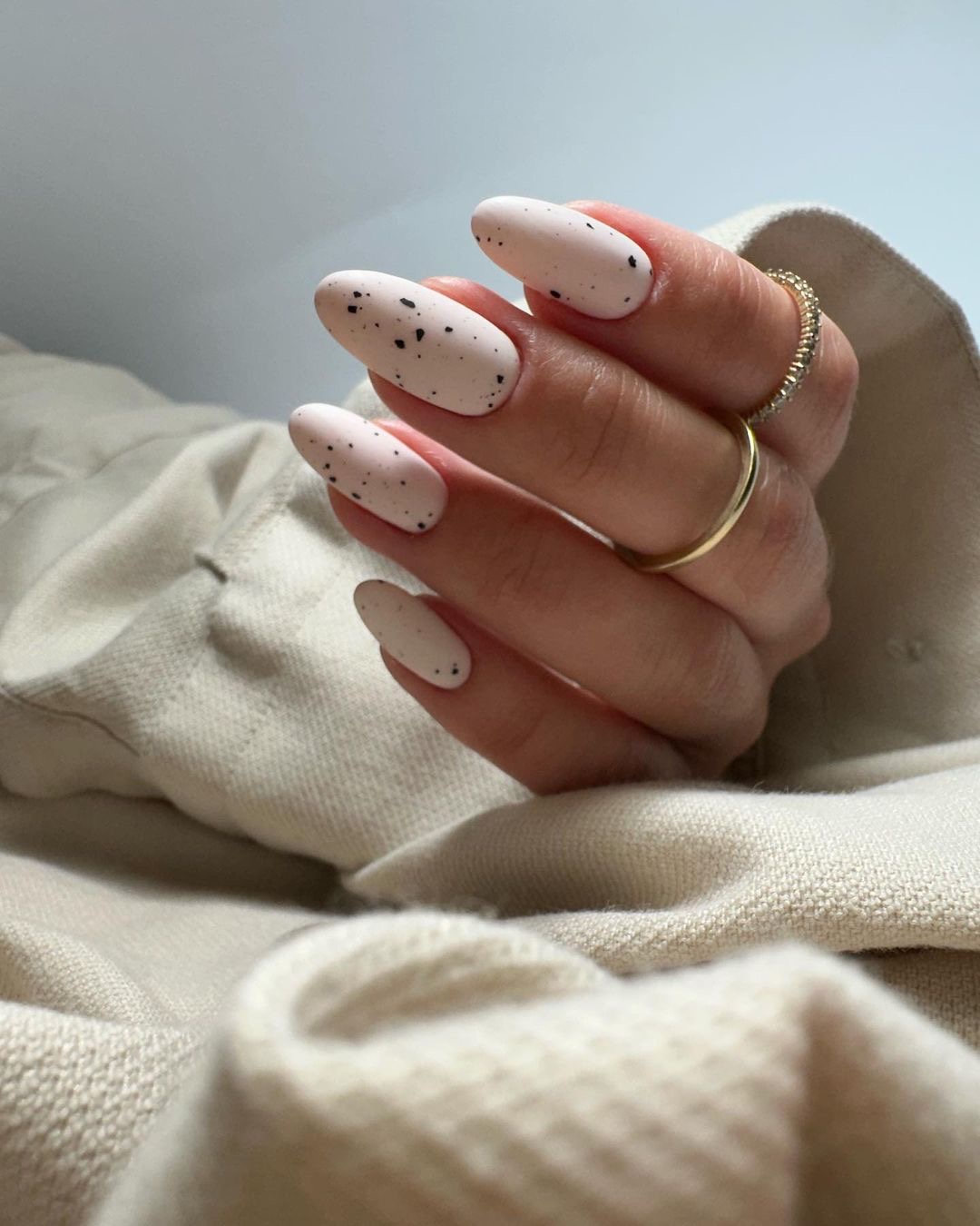 Dłoń z owalnymi paznokciami w odcieniu złamanej bieli z czarnymi plamkami, ozdobiona złotymi pierścionkami na tle beżowej tkaniny.
