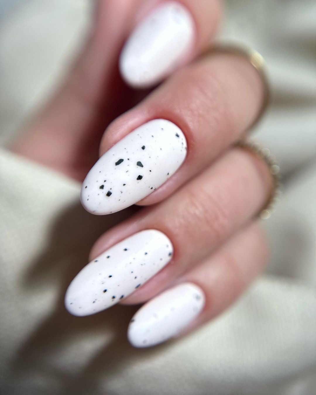 Dłoń z owalnymi paznokciami pokrytymi białym lakierem z czarnymi, nieregularnymi kropkami, na serdecznym palcu złoty pierścionek, tło kremowe.