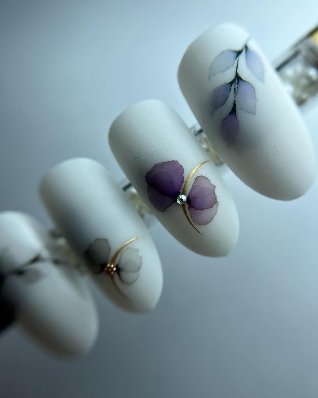 Delikatny, matowy manicure z motywem akwarelowych kwiatów w odcieniach fioletu i szarości, ozdobiony drobnymi, błyszczącymi cyrkoniami i cienkimi złotymi liniami.
