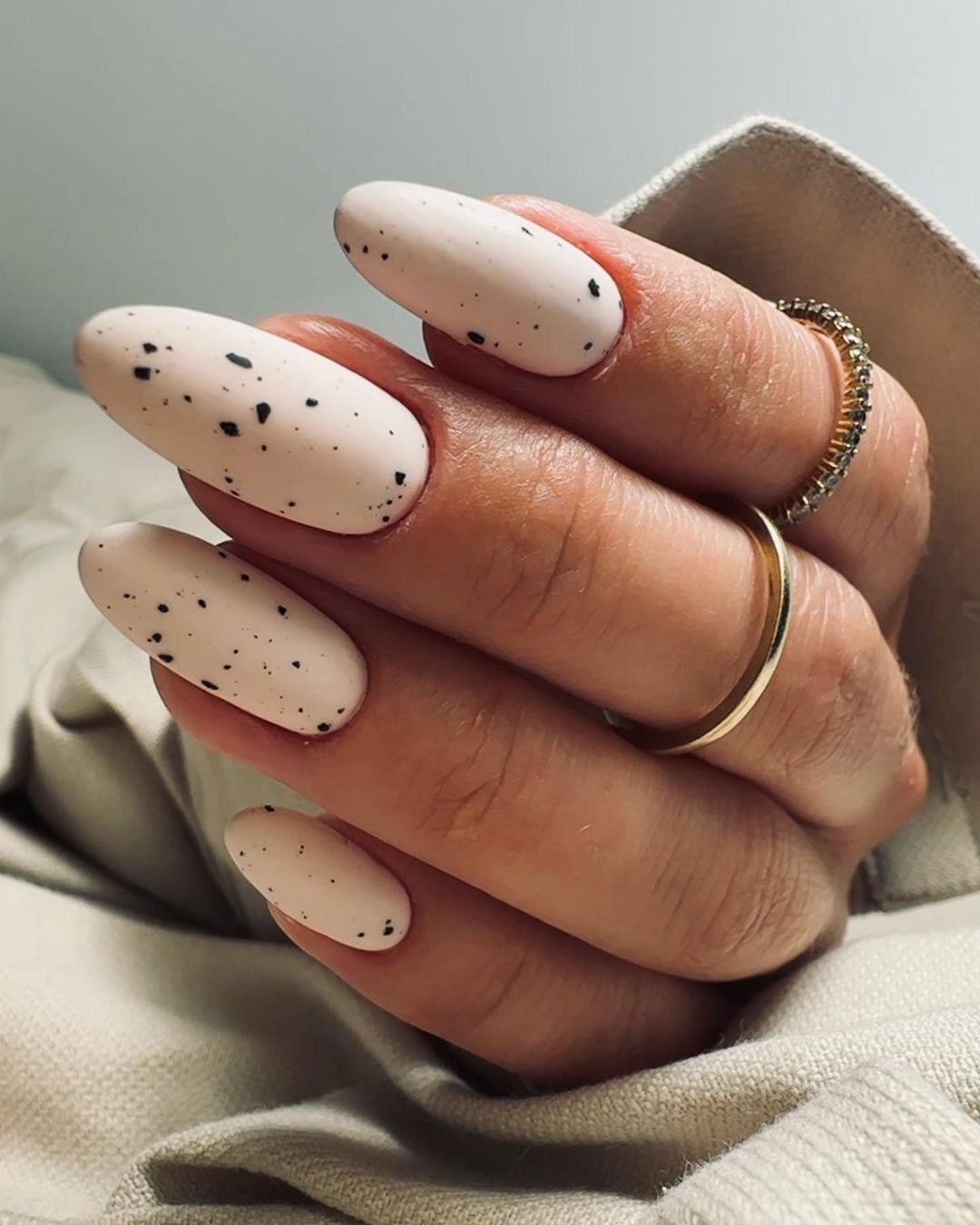 Paznokcie w kształcie migdała, pokryte jasnoróżowym lakierem z czarnymi kropkami, ozdobione złotymi pierścionkami na palcach.