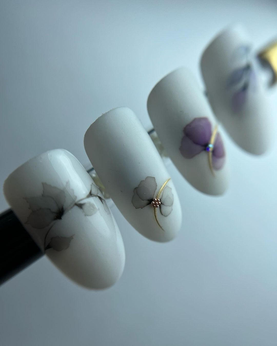 Cztery białe paznokcie z matowym wykończeniem, zdobione motywami kwiatowymi w odcieniach szarości i fioletu, z delikatnymi złotymi akcentami.