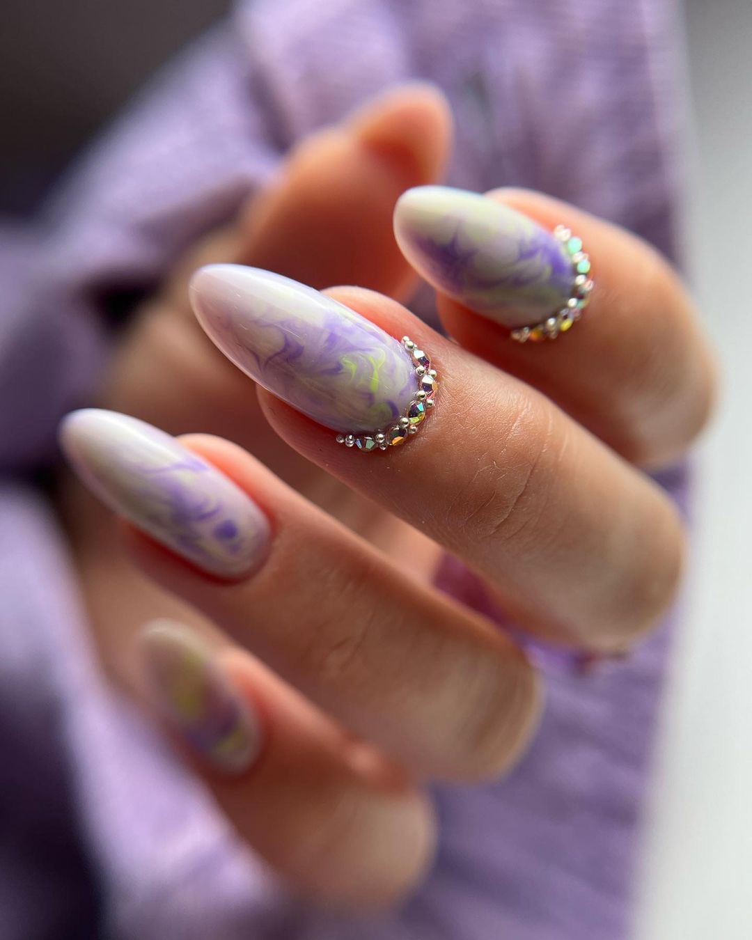 Paznokcie w odcieniach fioletu i bieli z efektem marmuru, ozdobione drobnymi kryształkami przy nasadzie, na tle liliowej tkaniny.