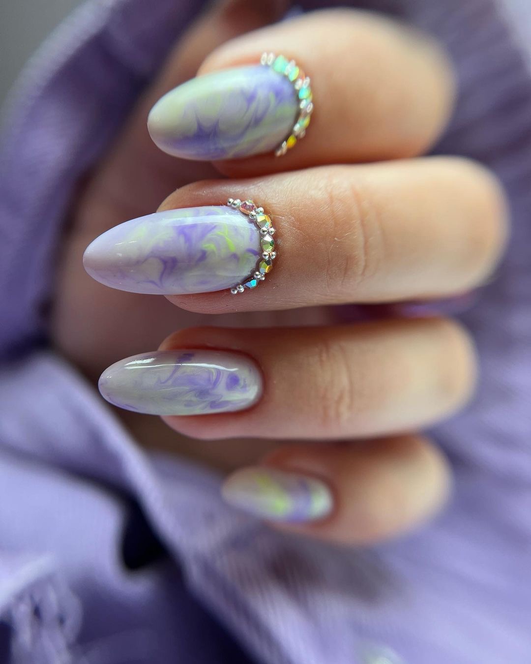 Delikatne, marmurkowe paznokcie w odcieniach lilii i zieleni, ozdobione rzędem drobnych, błyszczących kryształków przy skórkach, na tle fioletowej tkaniny.