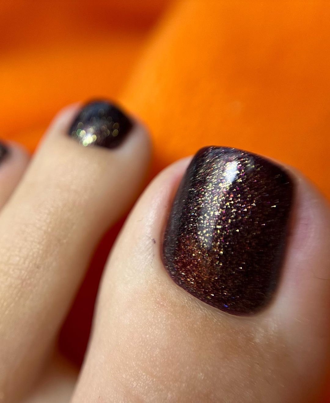 Zbliżenie na pomalowane paznokcie u stóp w kolorze ciemnego brązu z efektem brokatu na pomarańczowym tle.