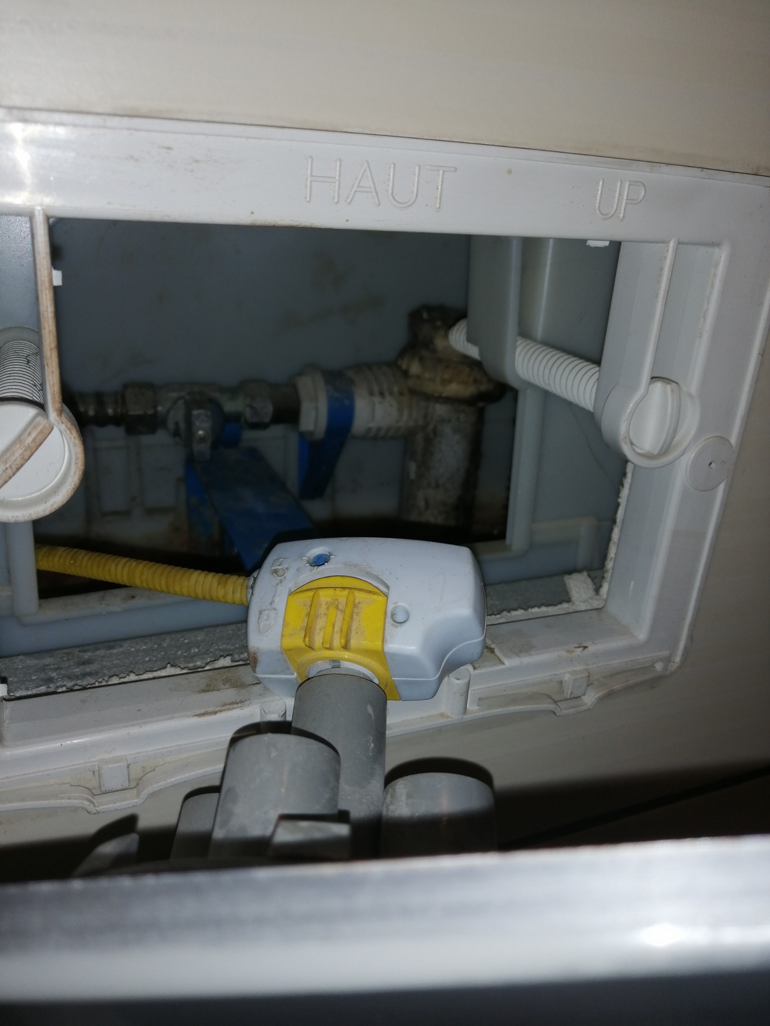 Wnętrze spłuczki toalety z widocznym zaworem, rurami i elementami mocującymi, oświetlone od góry, z napisami 'HAUT' i 'UP' na obudowie.