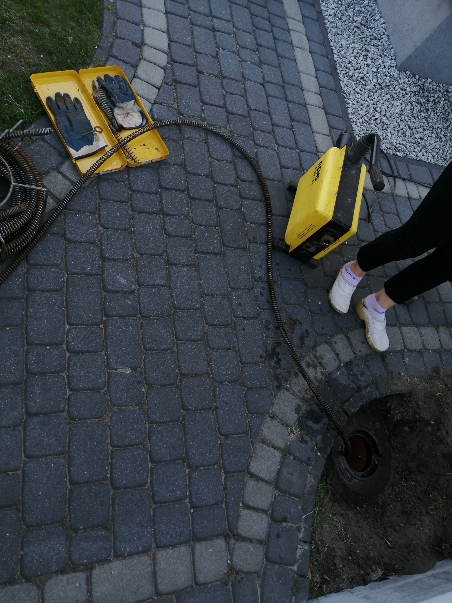 Żółta maszyna do udrażniania rur z wężem w kanalizacji na brukowanym podwórku, obok otwarte pudełko z roboczymi rękawicami.