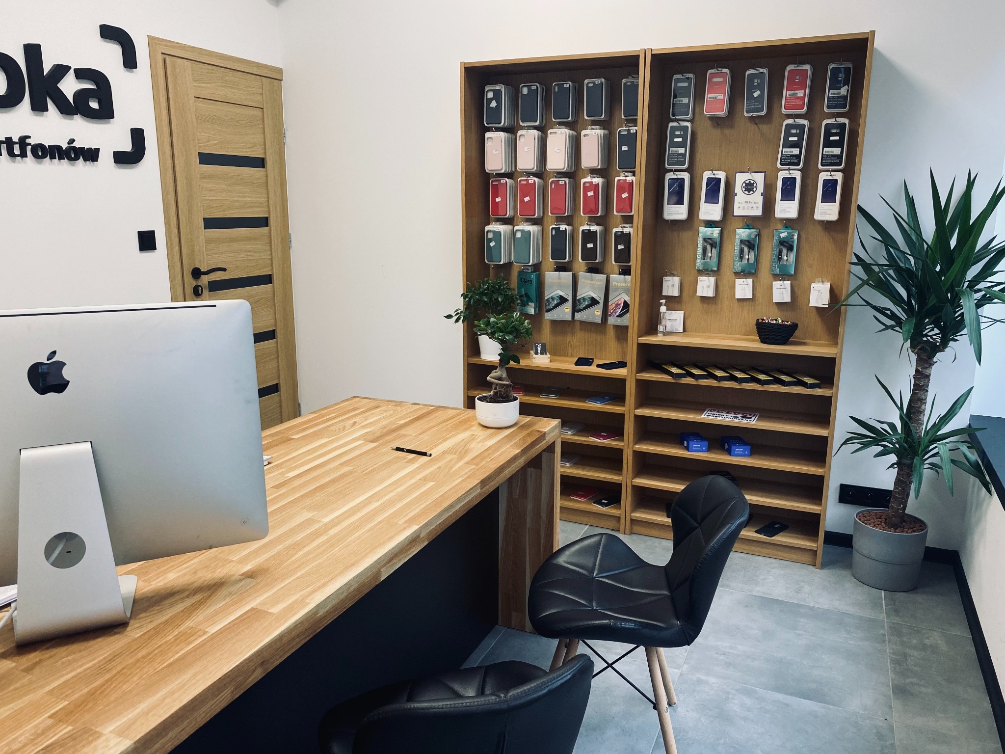 Wnętrze punktu obsługi z drewnianym biurkiem, komputerem Apple, dwoma czarnymi krzesłami i regałami z akcesoriami do telefonów, w tym etui i ładowarki.