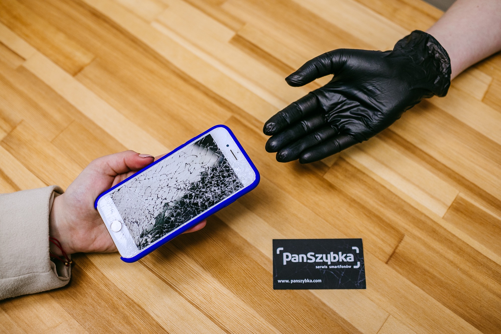 Dłoń w czarnej rękawiczce zbliża się do uszkodzonego smartfona z pękniętym ekranem w niebieskiej obudowie, leżącego na drewnianym blacie obok wizytówki serwisu.