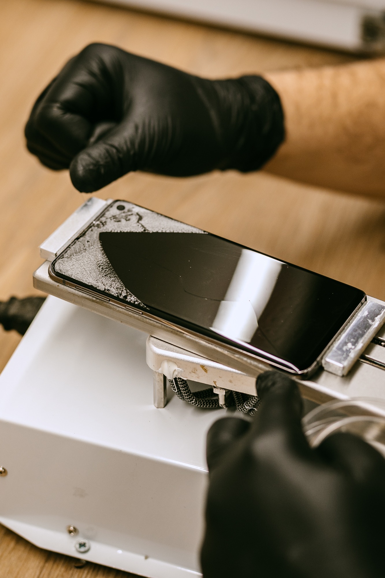 Smartfon z uszkodzonym ekranem umieszczony na maszynie serwisowej; dłonie w czarnych rękawiczkach przygotowują go do naprawy.