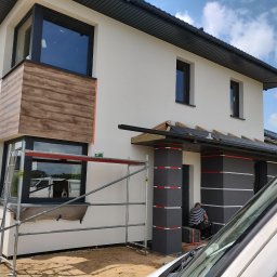 Polak-Bud - Ocieplanie elewacji domu jednorodzinnego z elementami dekoracyjnymi imitującymi drewno, widoczne rusztowanie i pracownik w trakcie montażu izolacji.