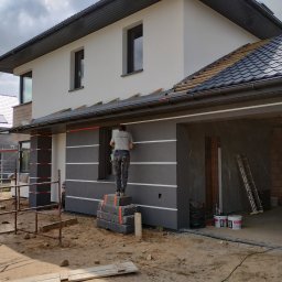 Polak-Bud - Pracownik na prowizorycznym podeście z betonowych bloków wykonuje prace na elewacji dwukondygnacyjnego domu z szarą elewacją z białymi poziomymi pasami, widoczne rusztowania i materiały budowlane.