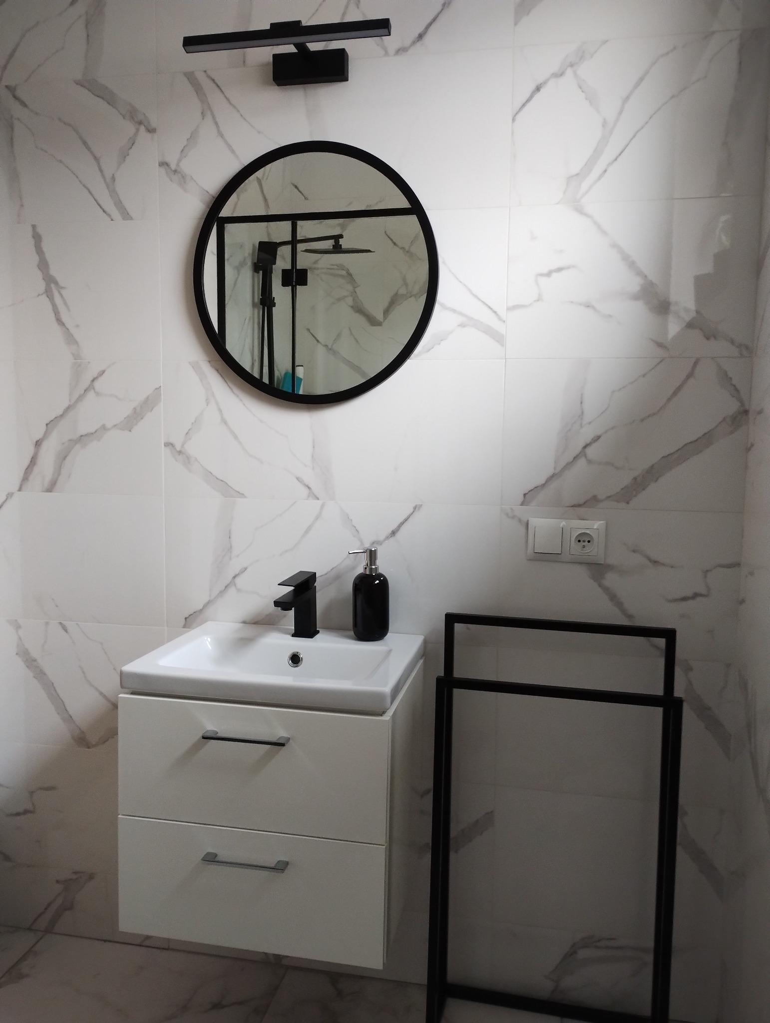 Nowoczesna łazienka z białą umywalką nablatową, czarną armaturą, okrągłym lustrem w czarnej ramie i marmurowymi płytkami na ścianach