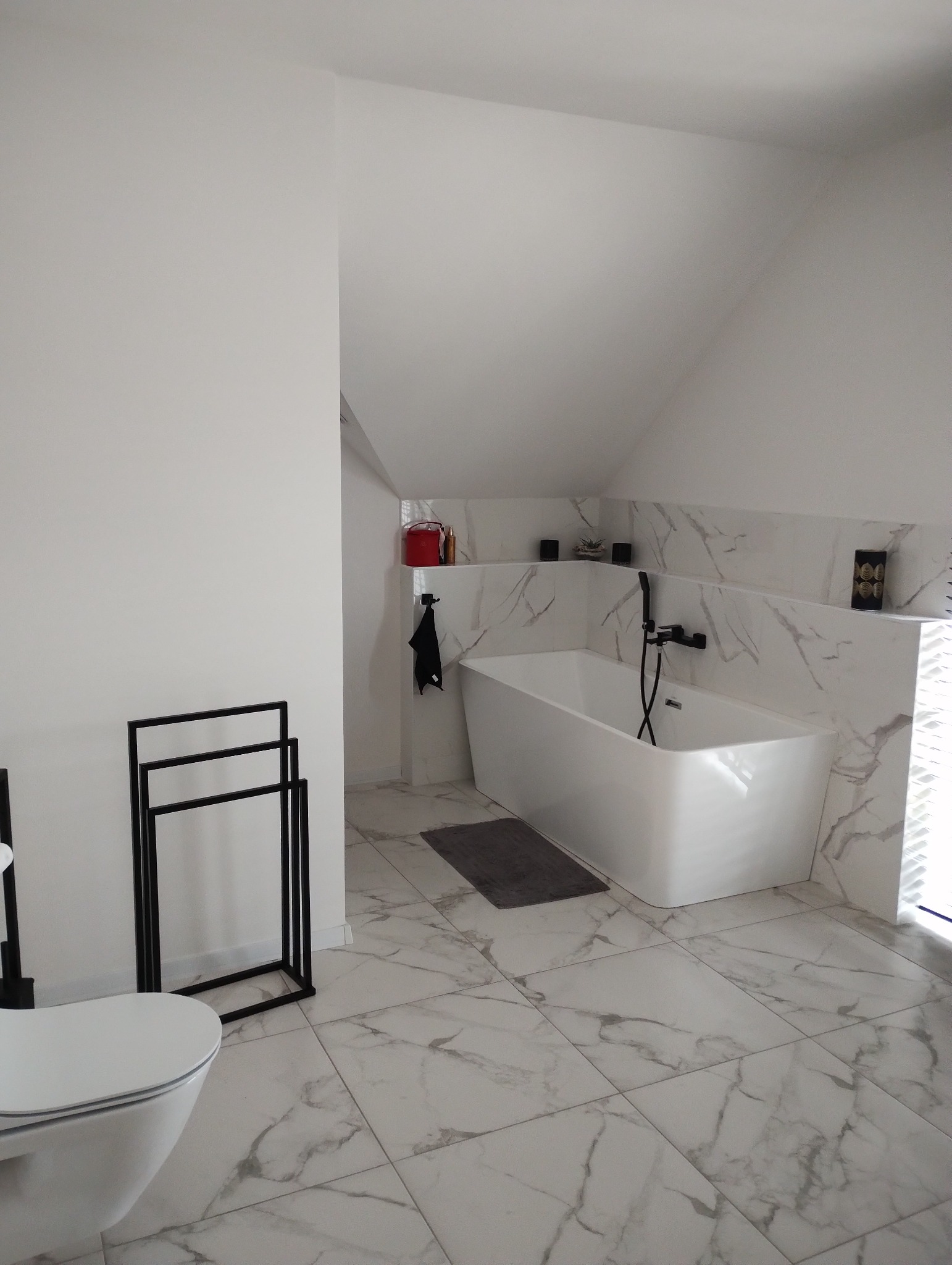 Nowoczesna łazienka na poddaszu z białą wolnostojącą wanną, marmurowymi płytkami i czarną armaturą, uzupełniona minimalistycznym wieszakiem na ręczniki i toaletą.