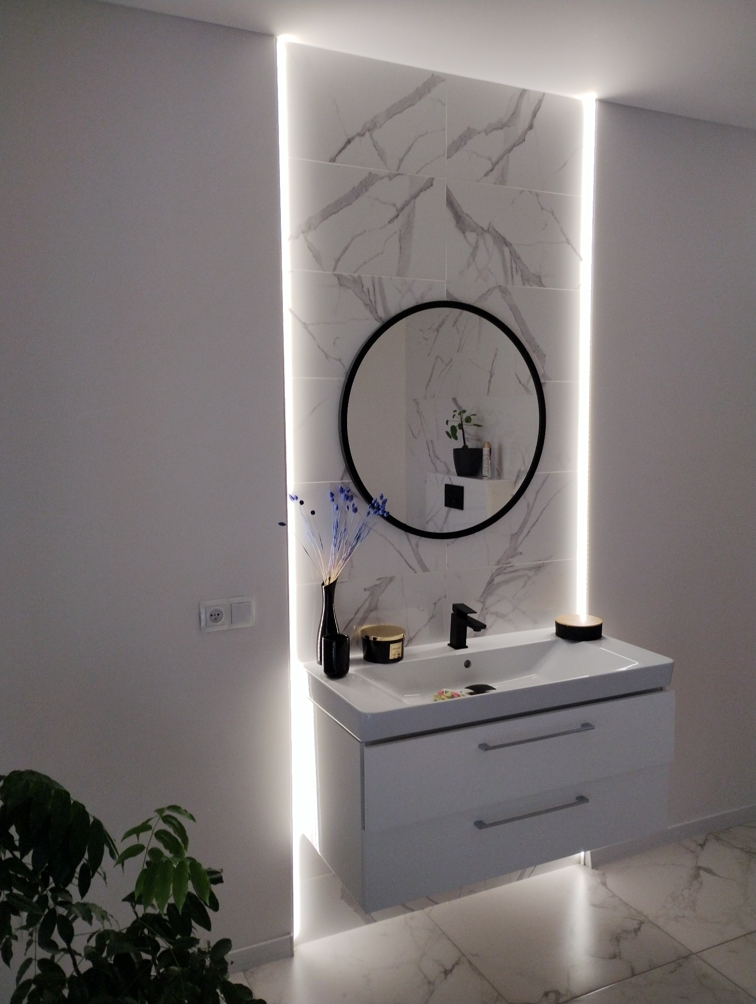 Nowoczesna łazienka z białymi marmurowymi płytkami, podświetleniem LED, okrągłym lustrem i białą szafką pod umywalką z czarną baterią.