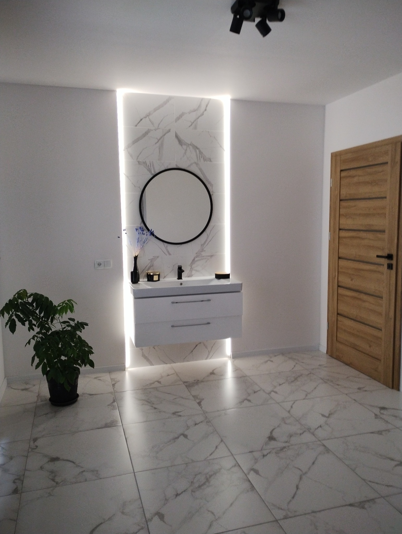 Nowoczesna łazienka z podświetlanym lustrem i marmurowymi płytkami podłogowymi, drewniane drzwi po prawej stronie, roślina w doniczce po lewej.