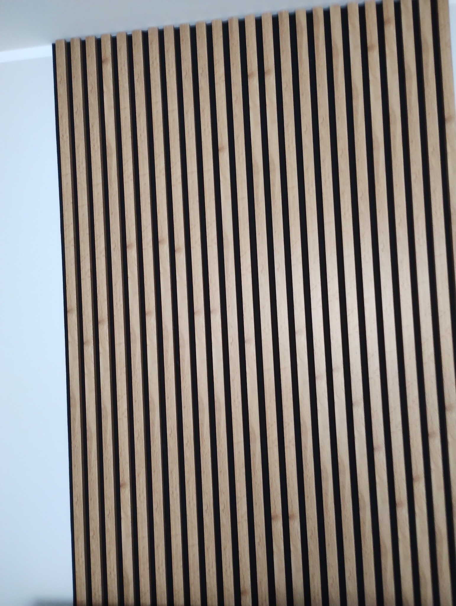 Drewniane lamele pionowe na czarnym tle, zamontowane na ścianie, widoczne z boku, tworzące dekoracyjny panel ścienny.