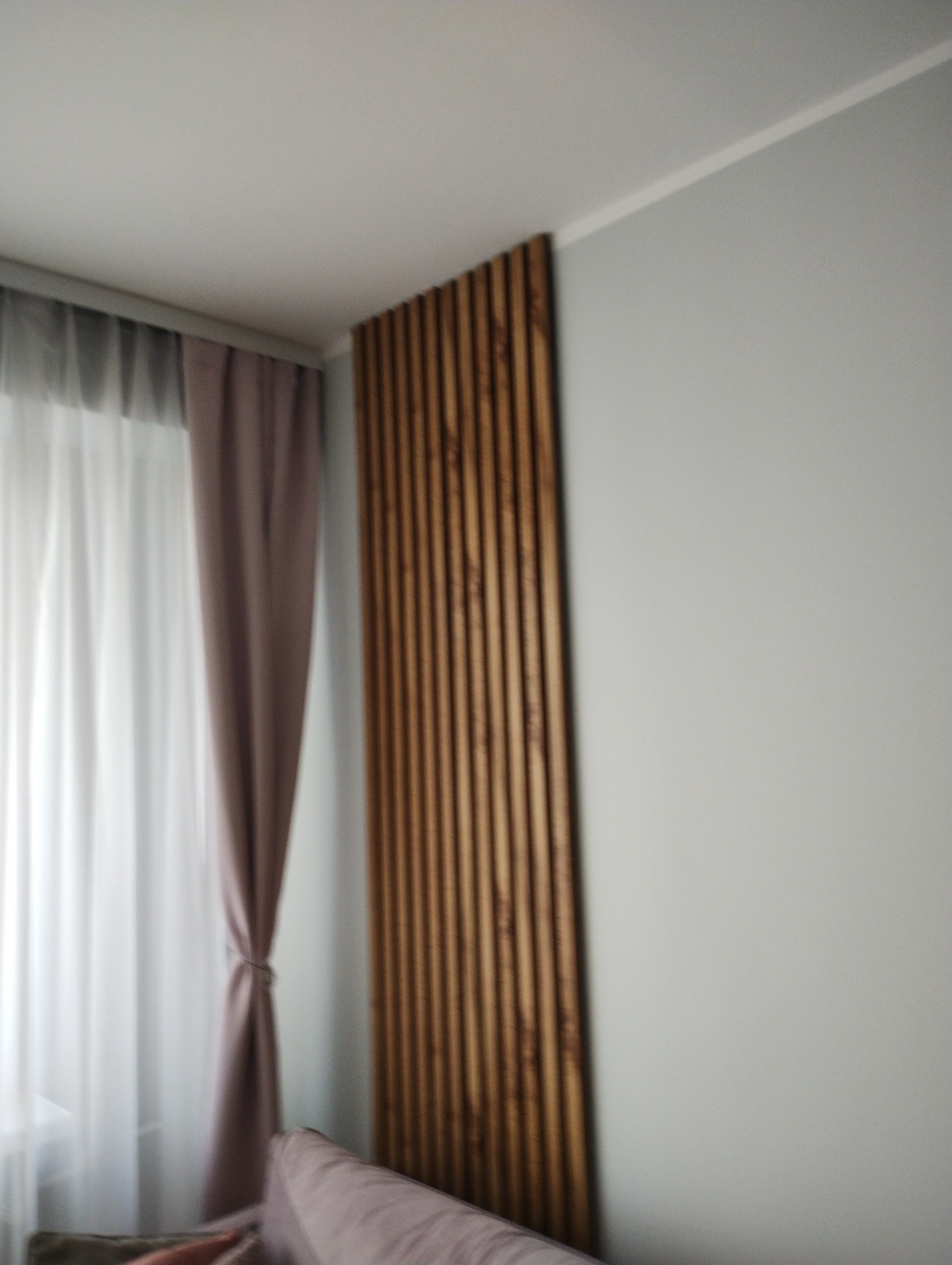 Drewniane lamele dekoracyjne na ścianie w pokoju, obok okna z zasłonami w odcieniach bieli i fioletu, widok z dołu.