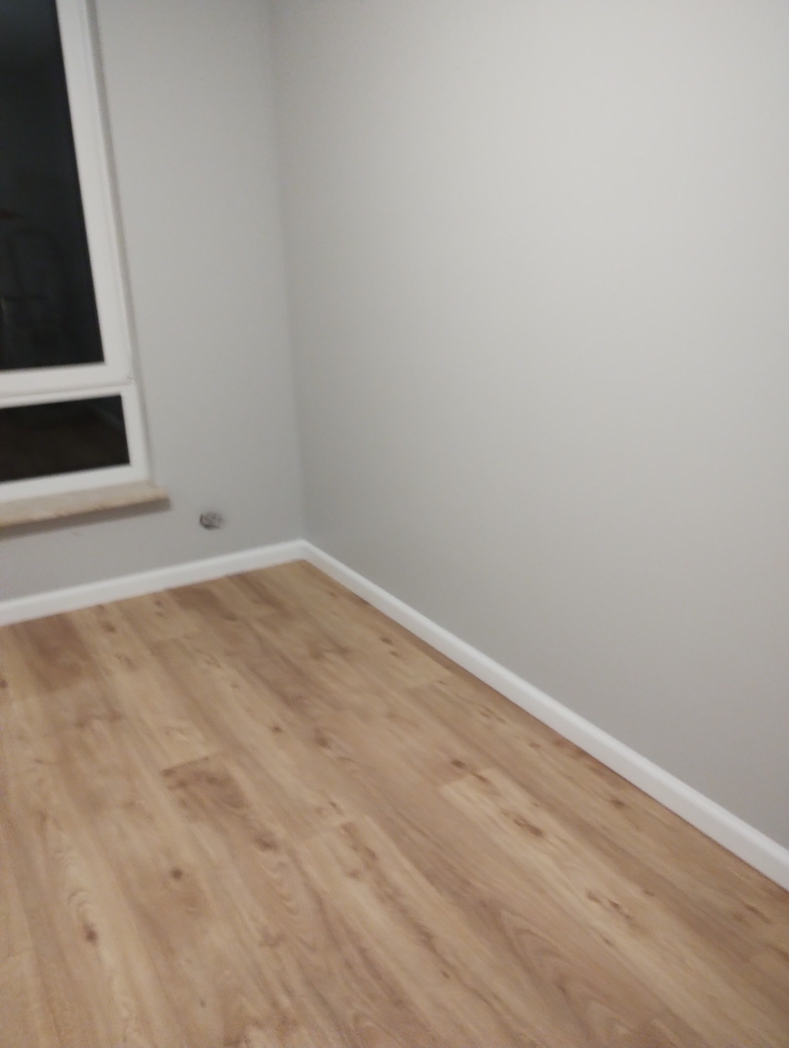 Wykończony narożnik pokoju z panelami podłogowymi w odcieniach brązu i szarą ścianą z oknem, widoczna listwa przypodłogowa.