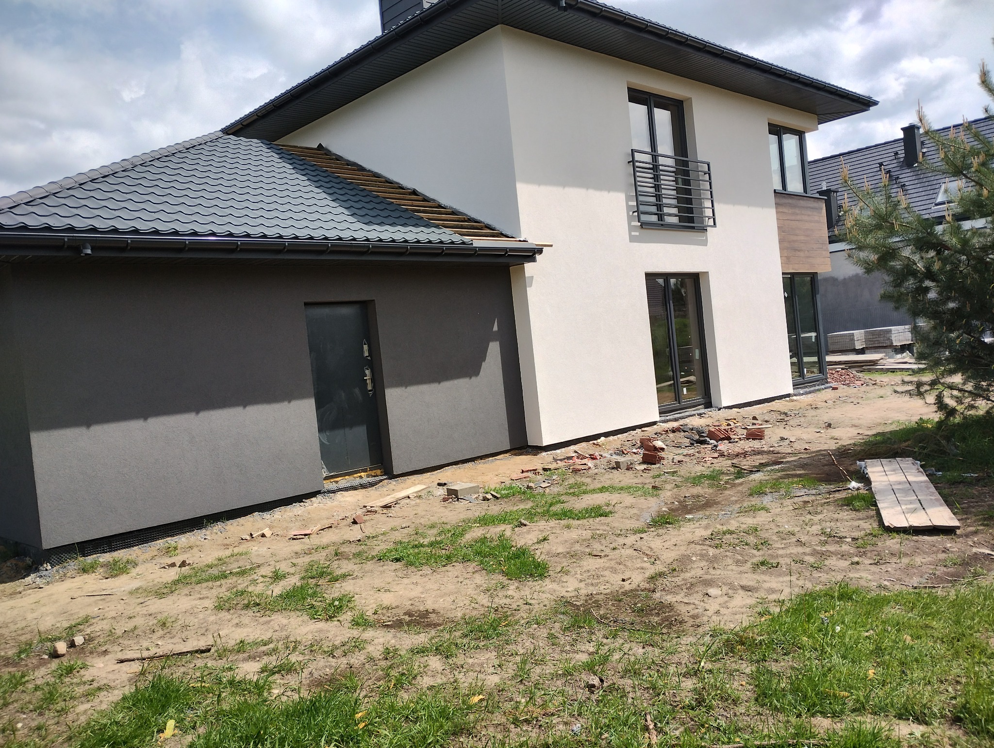 Dwukondygnacyjny dom jednorodzinny z szarą elewacją garażu i częściowo ocieploną elewacją frontową, widoczne porozrzucane cegły i deski na trawniku.