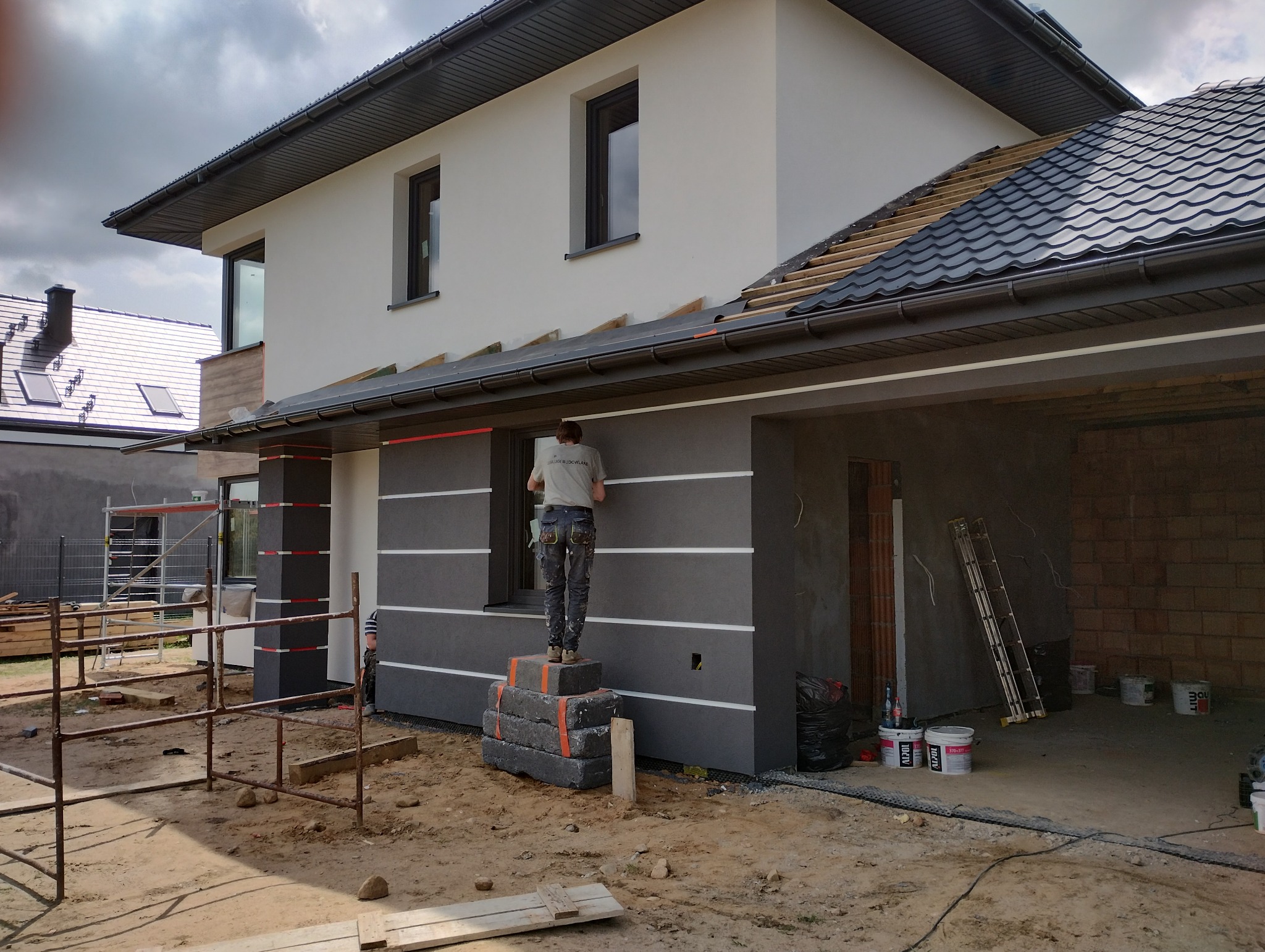 Pracownik na prowizorycznym podeście z betonowych bloków wykonuje prace na elewacji dwukondygnacyjnego domu z szarą elewacją z białymi poziomymi pasami, widoczne rusztowania i materiały budowlane.