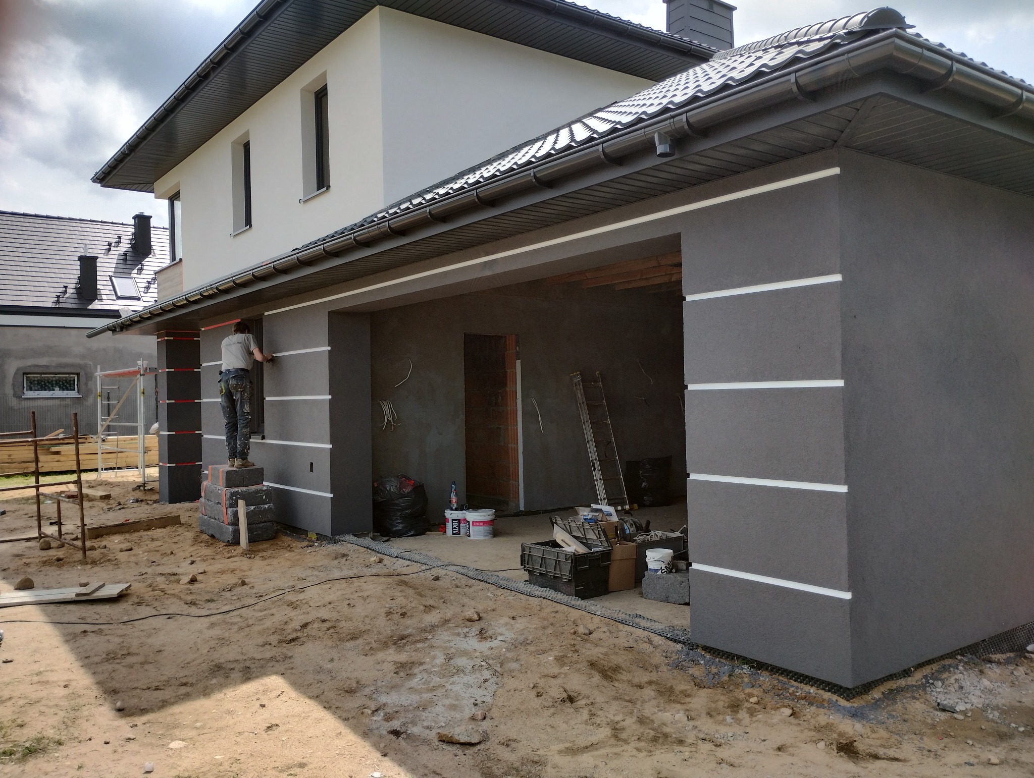 Pracownik na zewnątrz budynku, stojący na bloczkach, nakleja taśmę malarską na szarą elewację z białymi poziomymi paskami, przygotowując się do malowania.