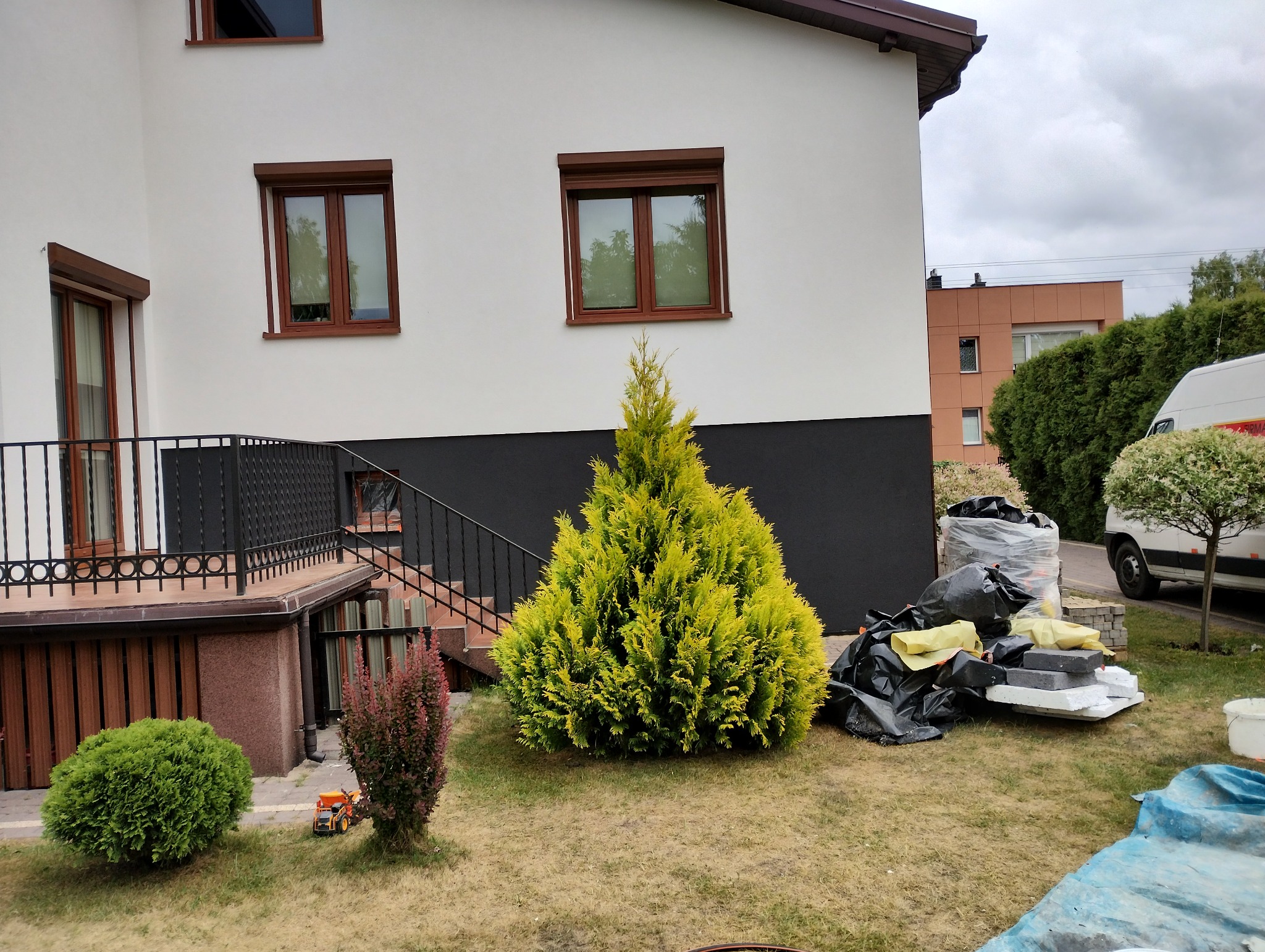 Biały dom z brązowymi oknami i czarnym cokołem, na trawniku leżą worki z odpadami budowlanymi i styropianem, w tle zaparkowany biały van.