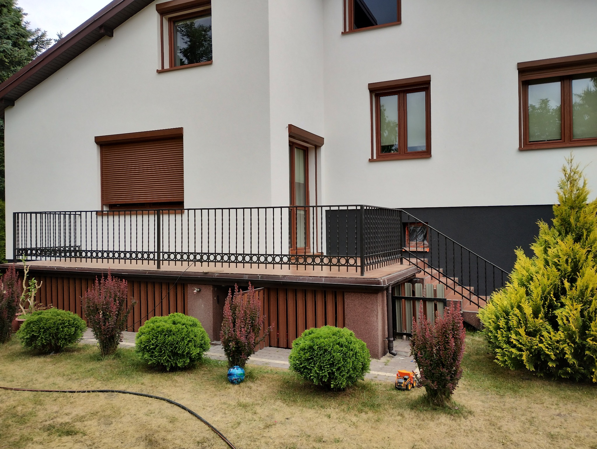 Jasna elewacja domu z brązowymi oknami i roletami, czarna balustrada balkonu i zadbany ogród z krzewami