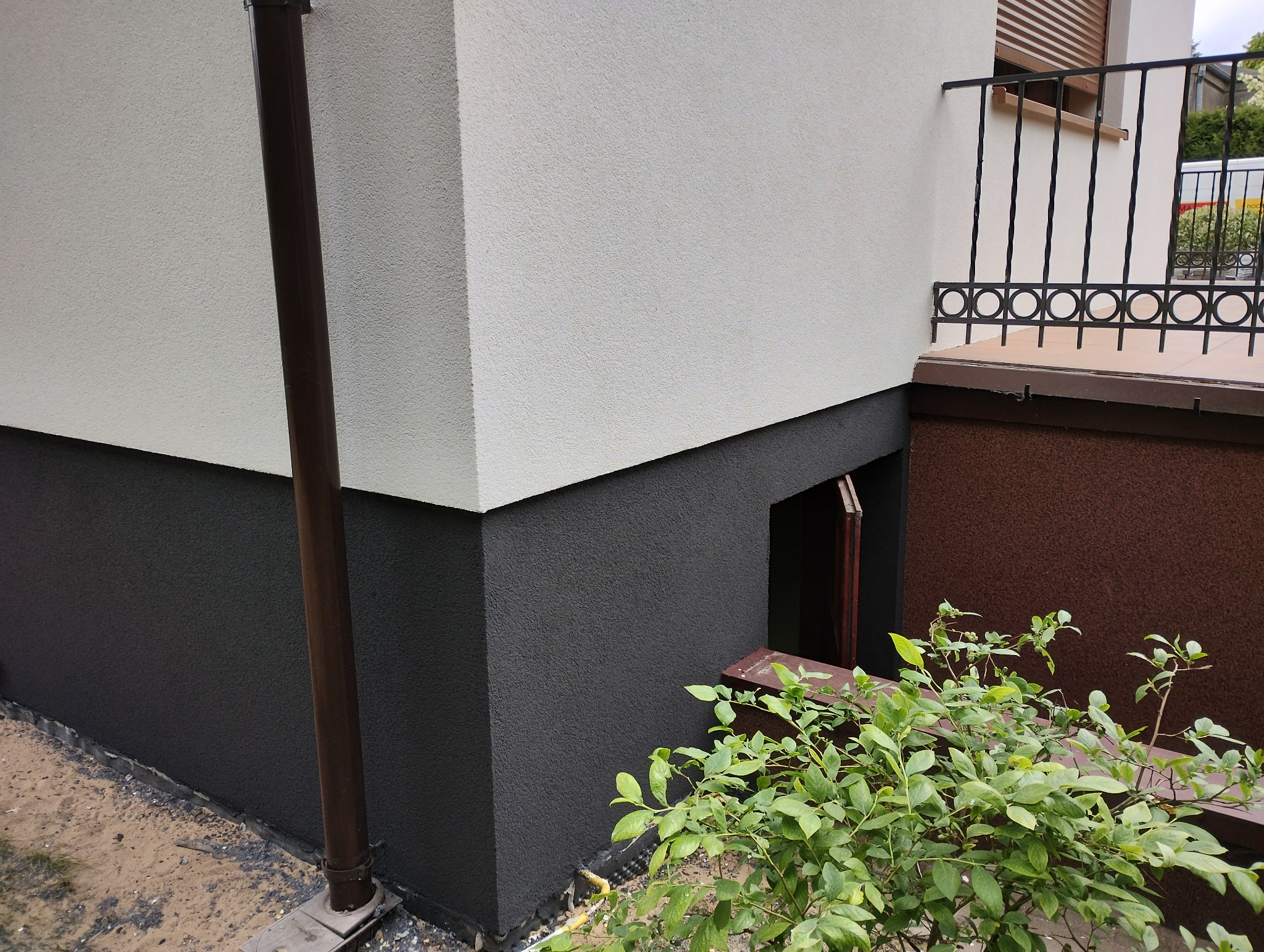 Dwukolorowa elewacja budynku z widocznym oknem piwnicznym, rynną i fragmentem balkonu.