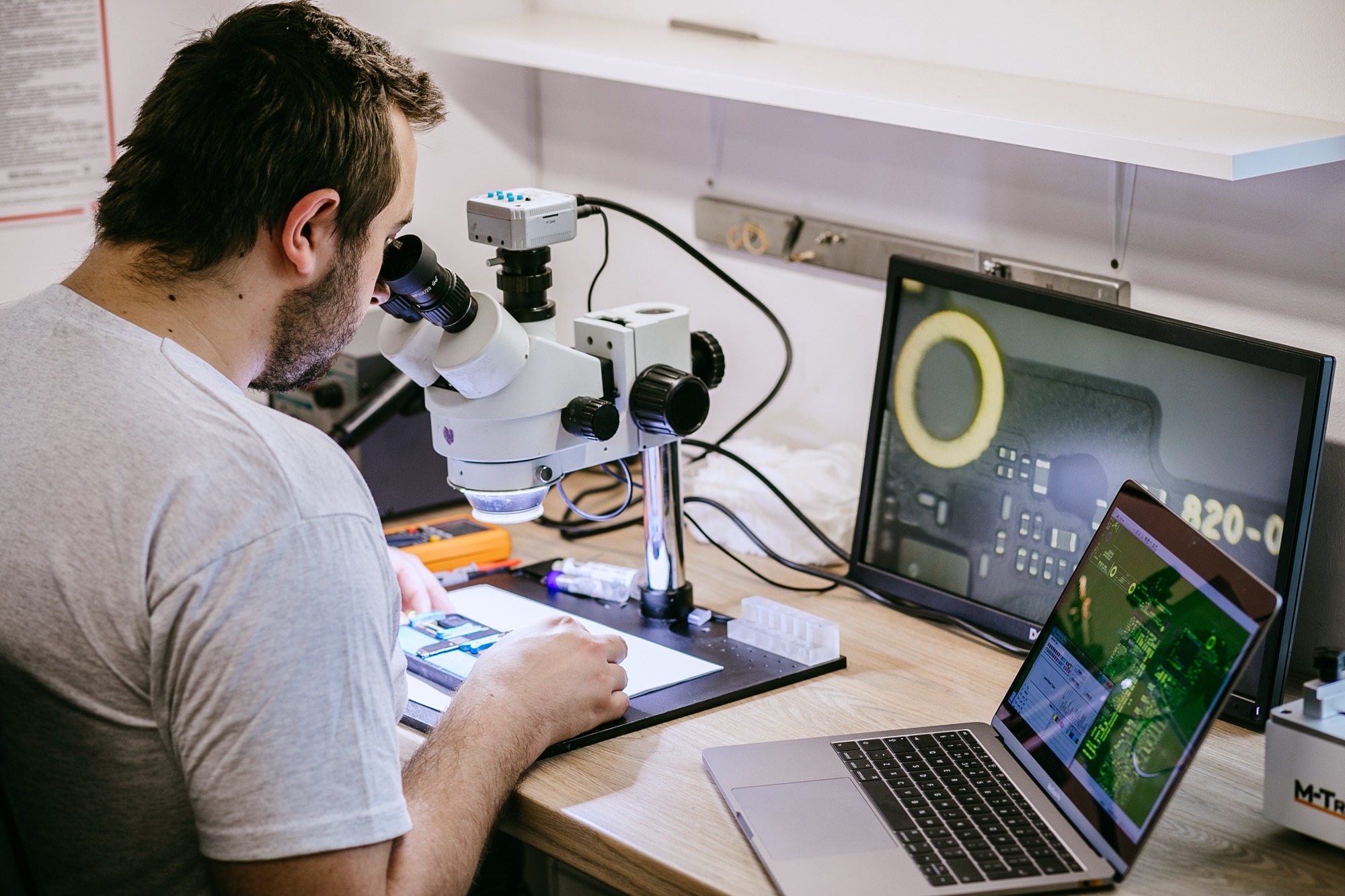 Technik pochylony nad mikroskopem, naprawiając płytę główną telefonu. Widoczny monitor z powiększonym obrazem elementu oraz laptop z schematem.