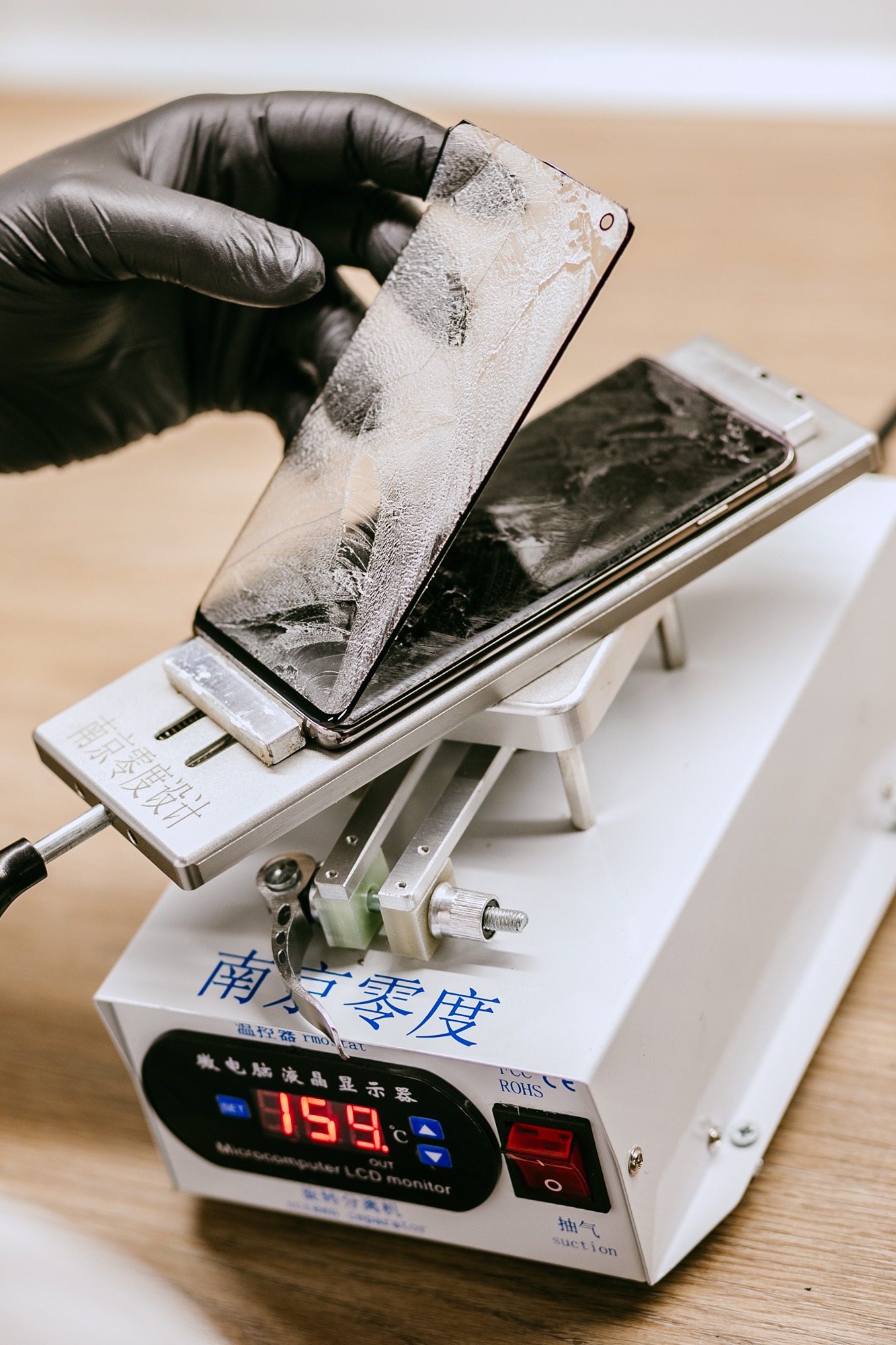 Uszkodzony wyświetlacz smartfona, trzymany w czarnej rękawicy, oddzielany od urządzenia na specjalistycznym stanowisku z wyświetlaczem temperatury.