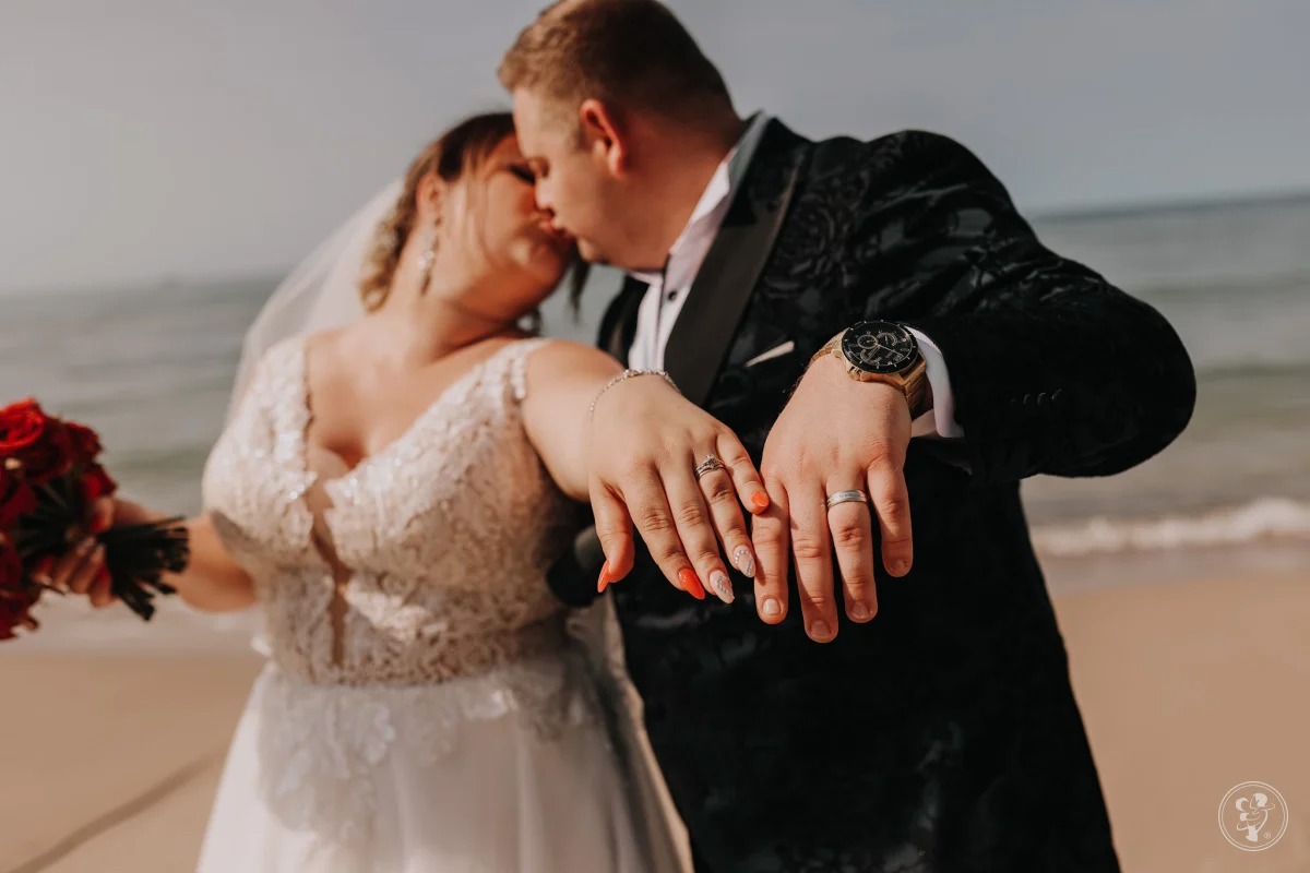 Para młoda na plaży pokazuje obrączki; panna młoda w koronkowej sukni trzyma bukiet czerwonych róż, pan młody w czarnym aksamitnym smokingu i złotym zegarku.