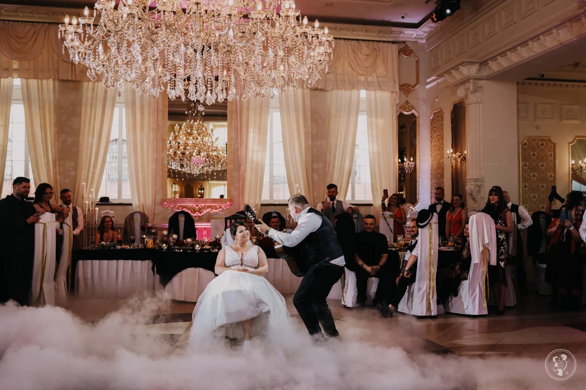 Panna młoda siedzi na krześle otoczona sztuczną mgłą, mężczyzna gra dla niej na gitarze, w tle elegancka sala balowa z żyrandolami i gośćmi.