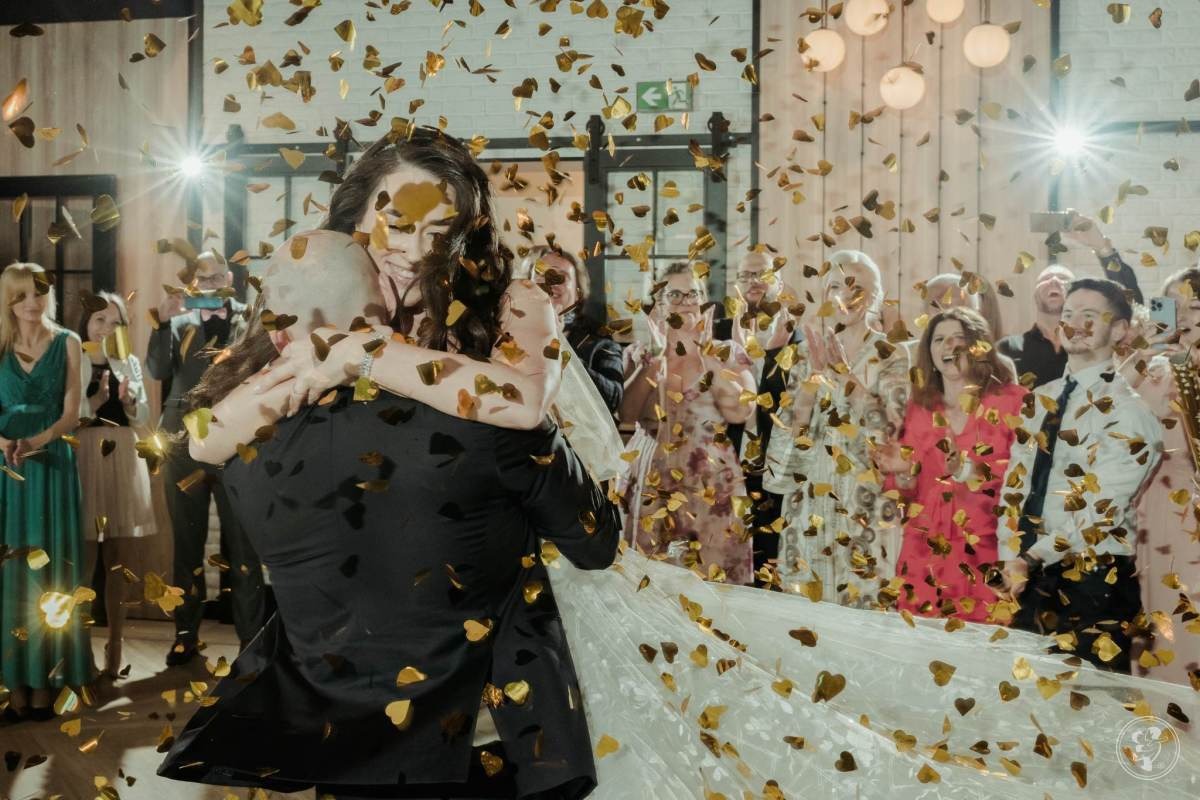 Para młoda w objęciach podczas pierwszego tańca, zasypana złotym konfetti w kształcie serc, w tle goście weselni i dekoracje sali.