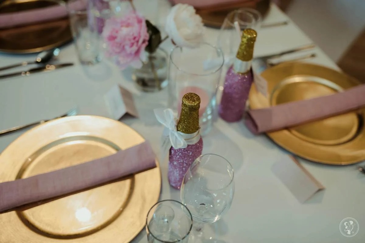 Elegancko nakryty stół z złotymi talerzami, fioletowymi serwetkami, kieliszkami i dekoracyjnymi buteleczkami z brokatem, kwiaty w wazonie.