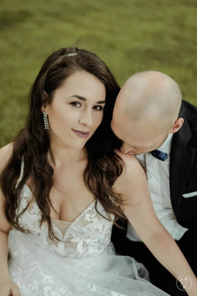 Panna młoda w koronkowej sukni spogląda w obiektyw, podczas gdy pan młody w ciemnym garniturze delikatnie całuje ją w szyję na tle zieleni.