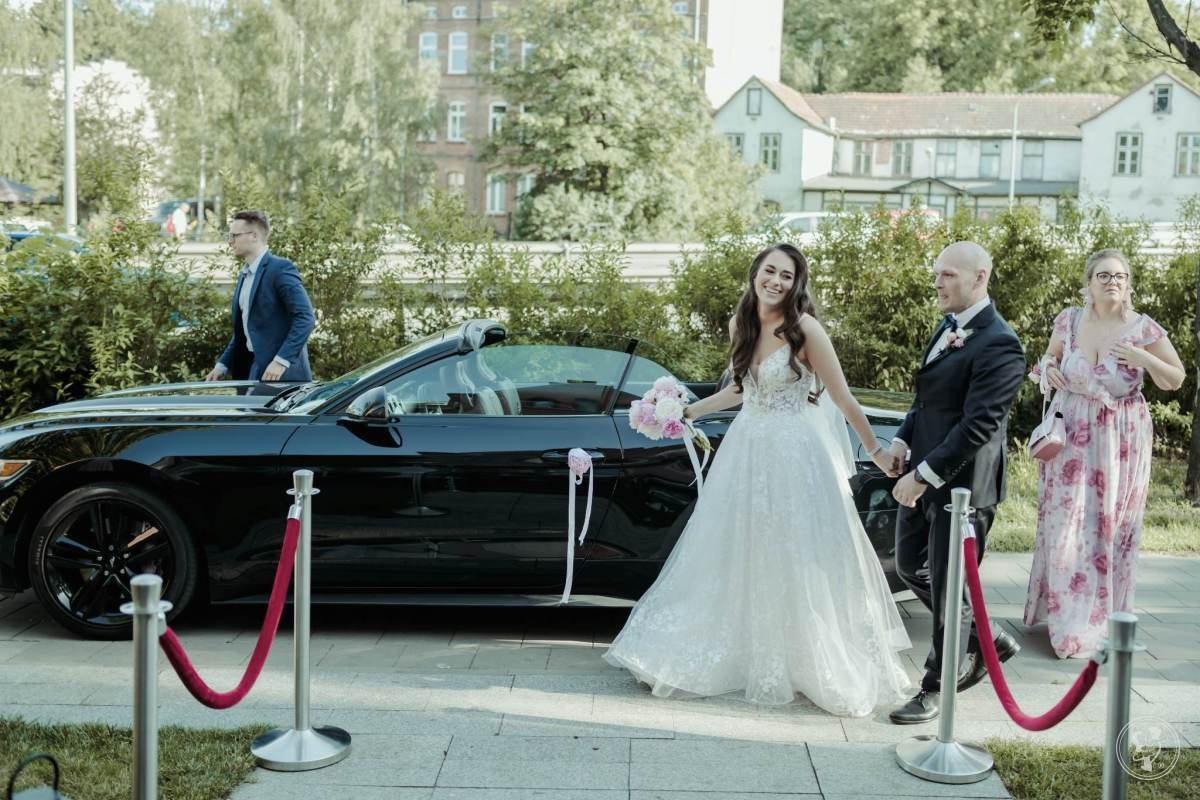 Uśmiechnięta panna młoda w koronkowej sukni wychodzi z czarnego kabrioletu Ford Mustang, trzymając bukiet różowych kwiatów, obok stoi pan młody w garniturze, za nimi kobieta w kwiecistej sukience...