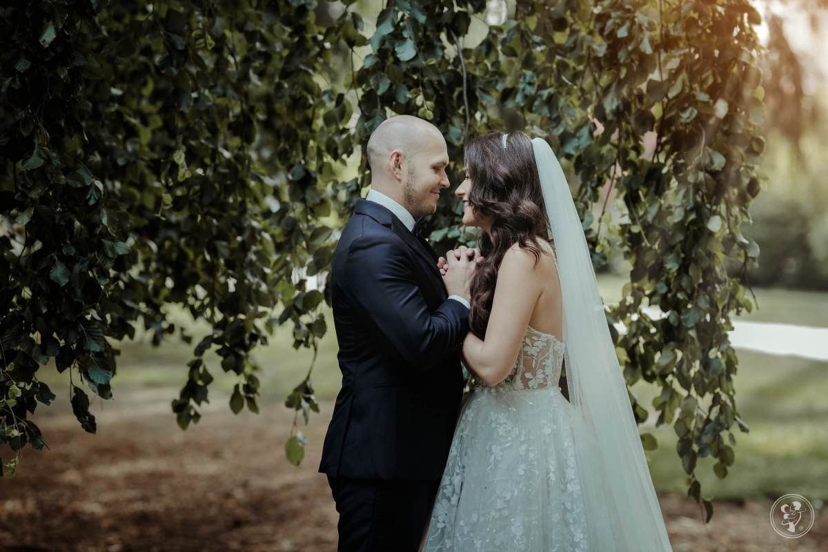 Para młoda, mężczyzna w granatowym garniturze i kobieta w koronkowej sukni ślubnej i welonie, stoją pod drzewem z opadającymi gałęziami, trzymając się za ręce i patrząc sobie w oczy.