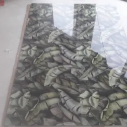 Ułożone płytki podłogowe z wzorem liści bananowca, odbijające światło, na jasnej posadzce, obok kartonowe opakowania i para brązowych but&oacute;w.