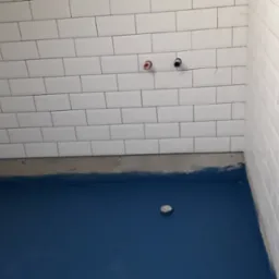 Niebieska hydroizolacja podłogi w łazience, białe kafelki ułożone do połowy ściany, widoczne rury instalacji wodnej, fragment nieotynkowanej ściany z ułożonymi płytkami.