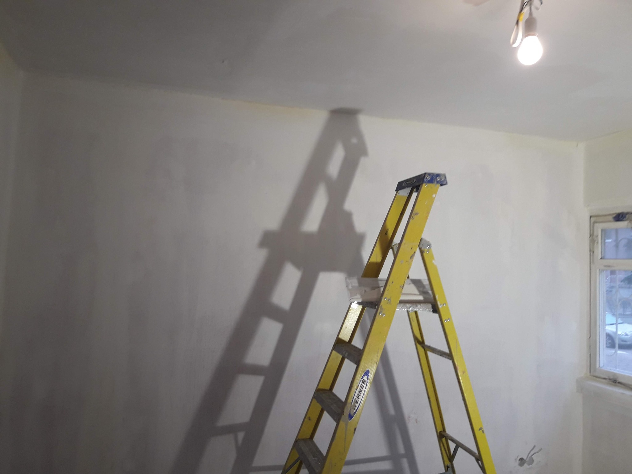 Żółta drabina rozstawiona w rogu pomieszczenia z oknem, ściany pokryte świeżo nałożoną, jasną warstwą, widoczny cień drabiny na ścianie, sufit z żarówką.