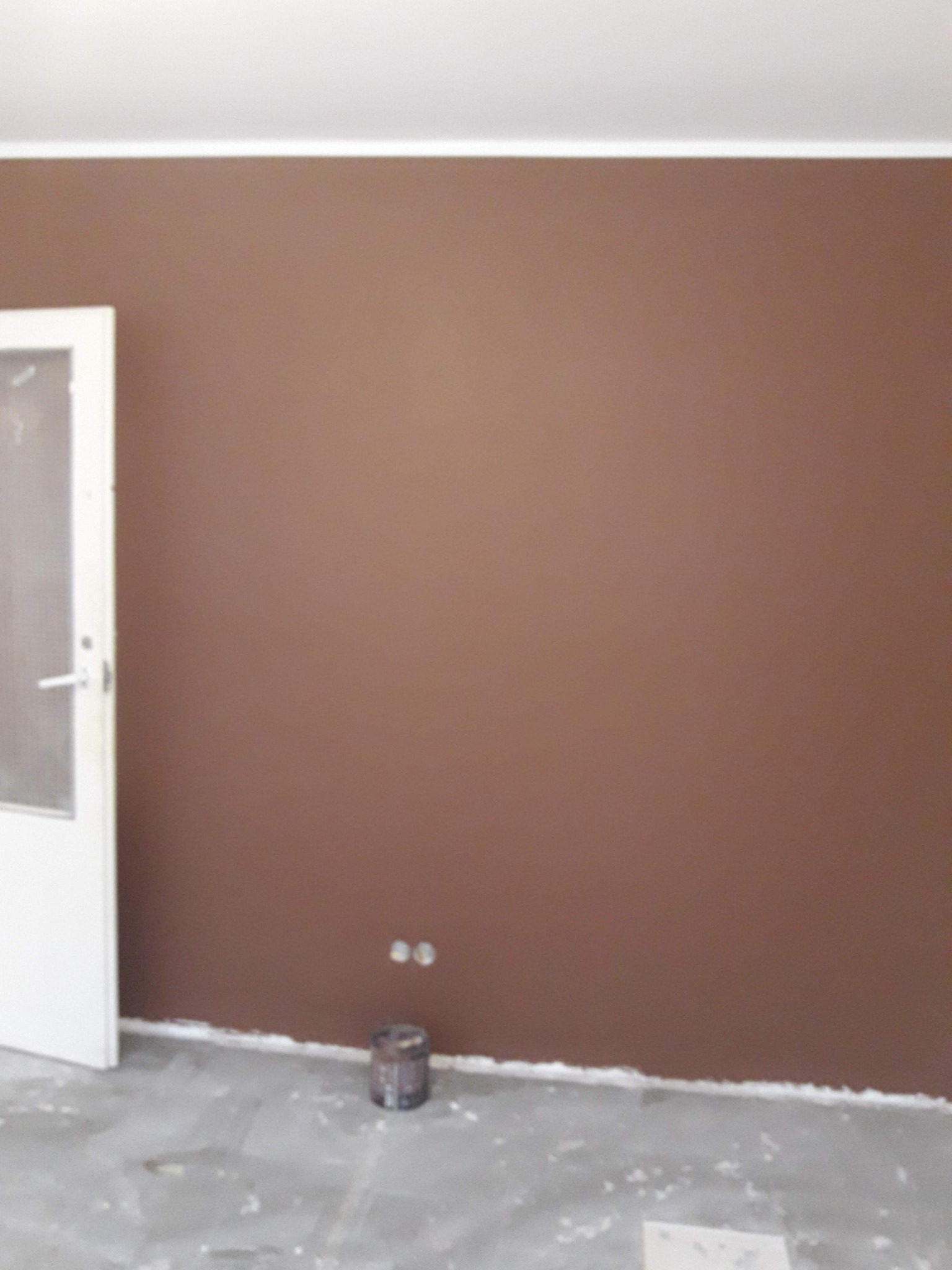 Świeżo pomalowana ściana w odcieniach brązu, widoczne dwa gniazdka elektryczne i puszka po farbie przy podłodze, białe listwy przypodłogowe i biała ościeżnica drzwi po lewej stronie.