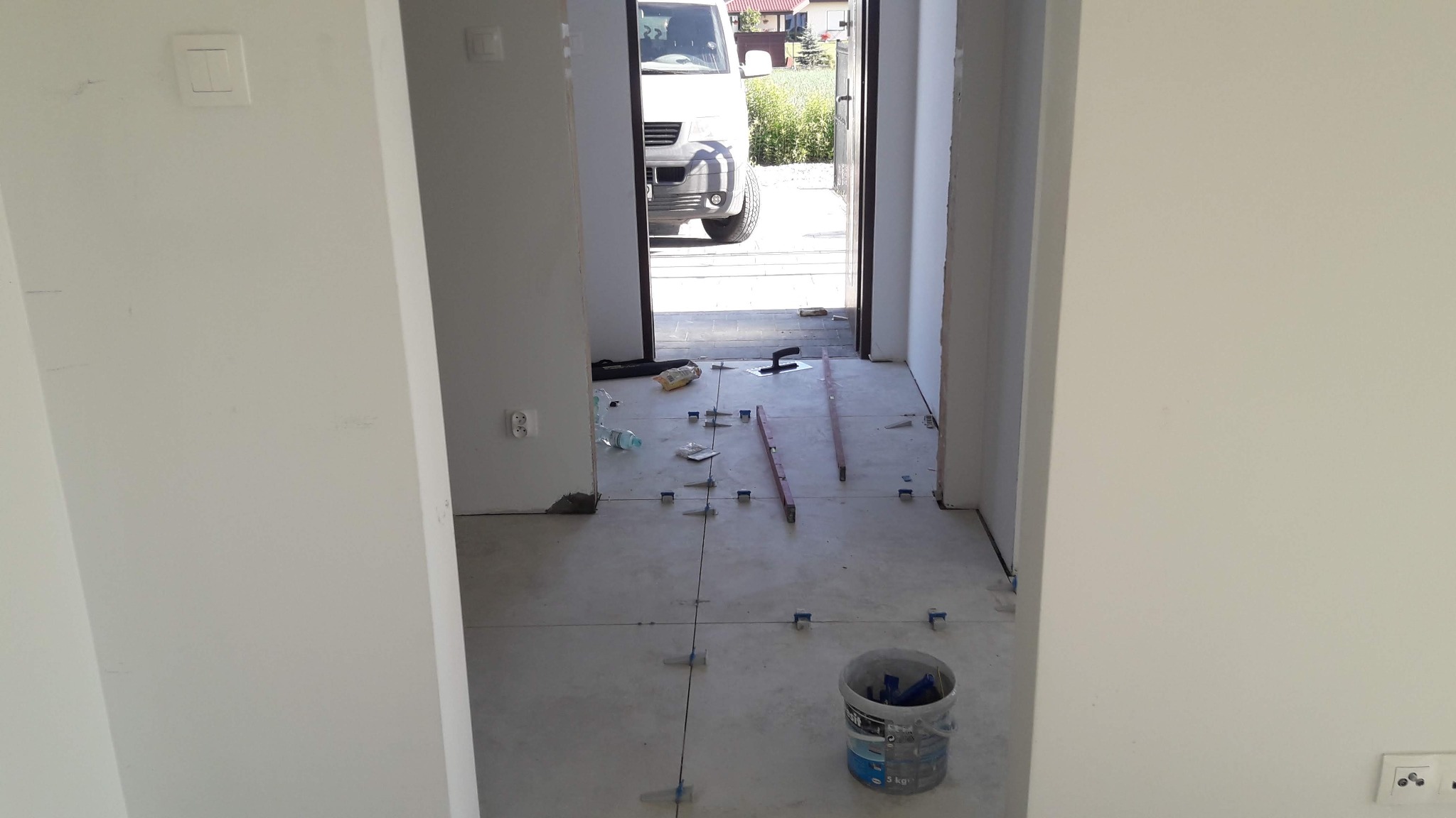 Układanie płytek podłogowych w korytarzu, widoczne poziomice, krzyżyki dystansowe, wiadro z klejem i zaparkowany bus za drzwiami.