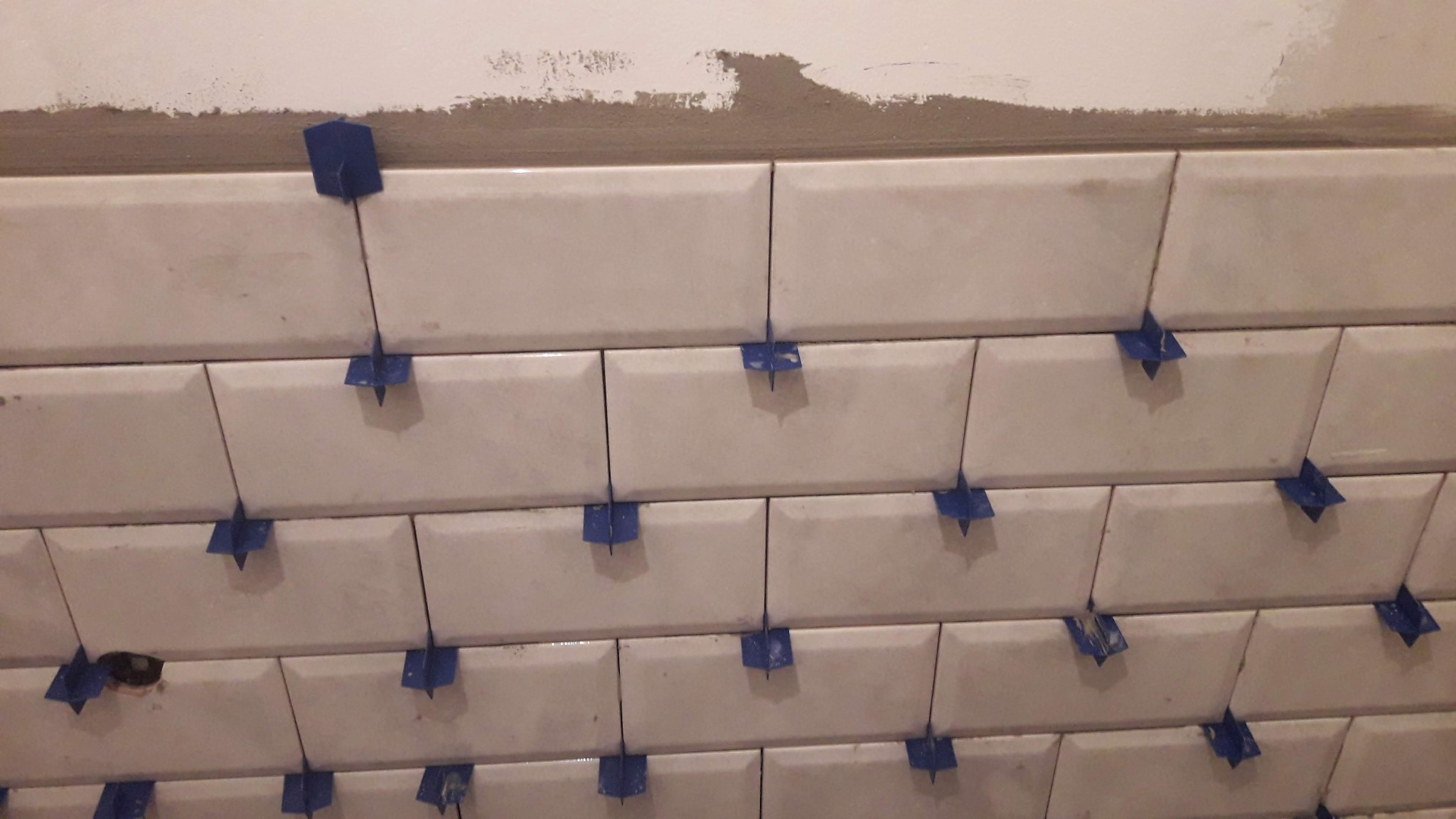 Układanie białych, prostokątnych płytek na ścianie z użyciem niebieskich krzyżyków dystansowych; widoczny fragment ściany z resztkami zaprawy.