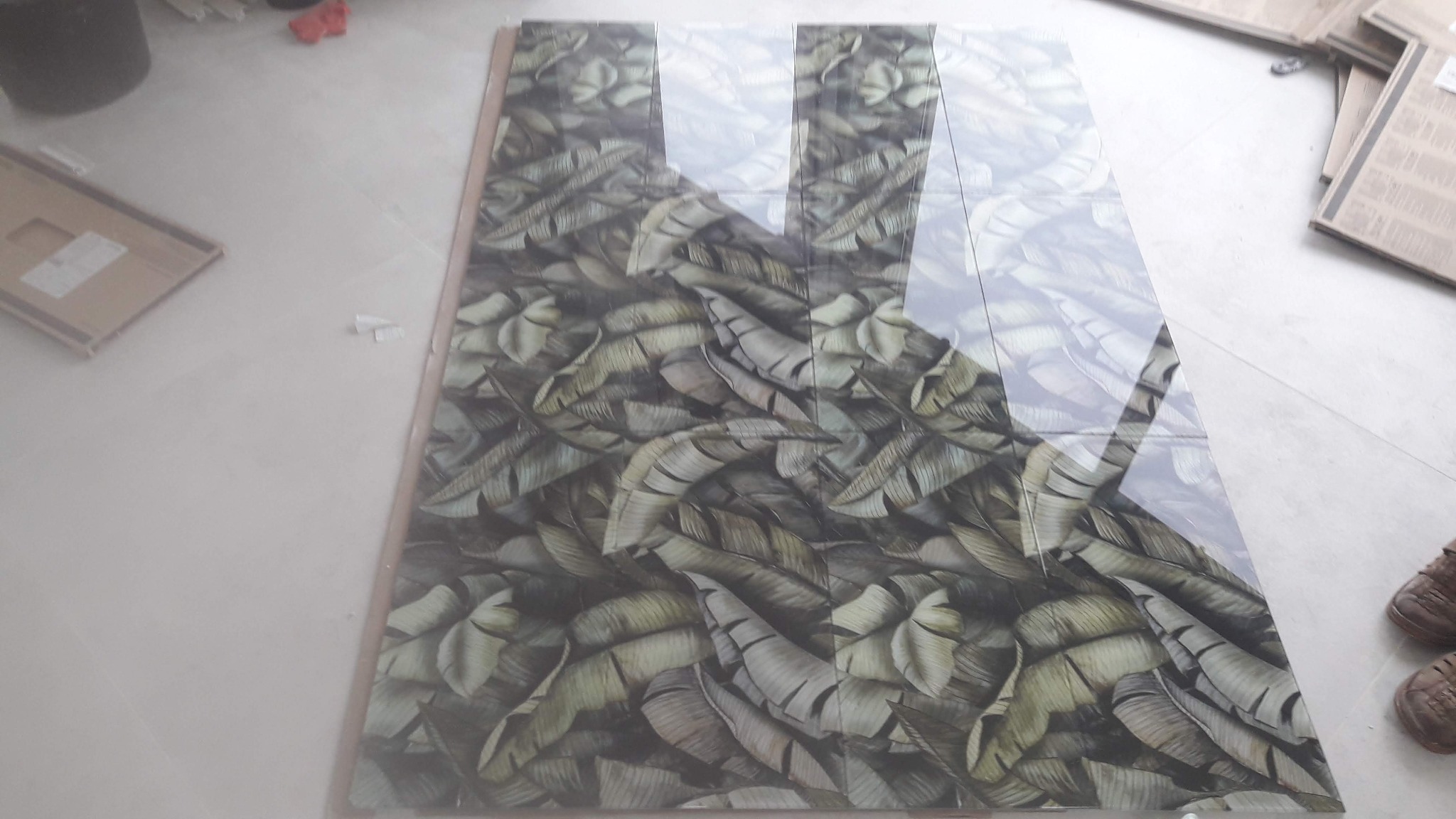 Ułożone płytki podłogowe z wzorem liści bananowca, odbijające światło, na jasnej posadzce, obok kartonowe opakowania i para brązowych butów.