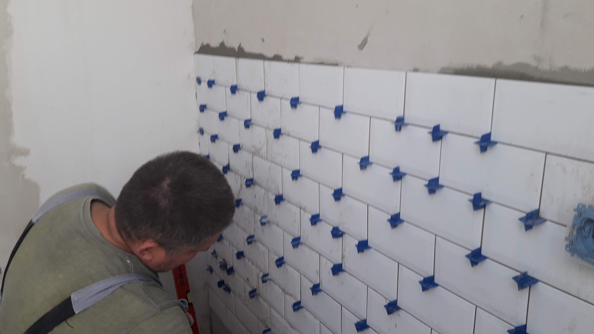 Układanie białych płytek na ścianie z użyciem niebieskich krzyżyków dystansowych, widoczny pracownik z poziomicą.