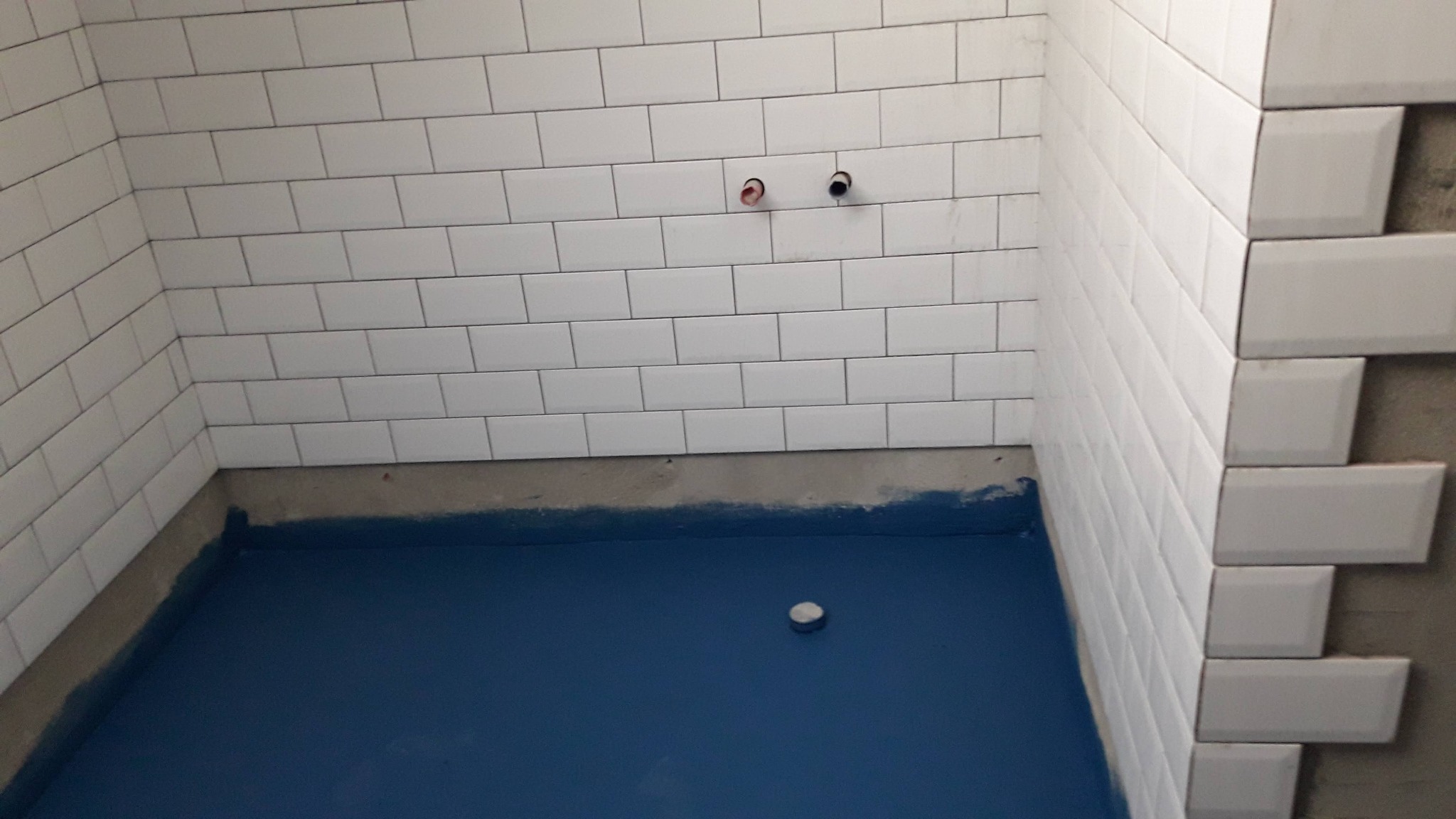 Niebieska hydroizolacja podłogi w łazience, białe kafelki ułożone do połowy ściany, widoczne rury instalacji wodnej, fragment nieotynkowanej ściany z ułożonymi płytkami.