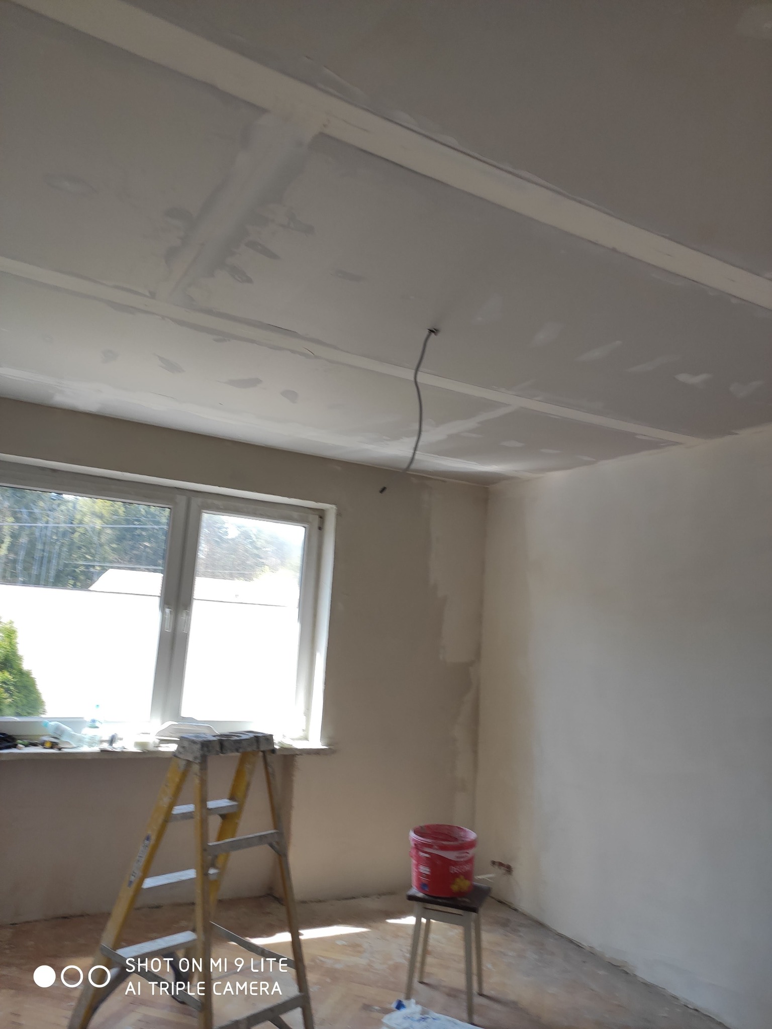 Pokój w trakcie remontu: gładzie na suficie i ścianach, drabina stoi przy oknie, wiadro z farbą na stołku.