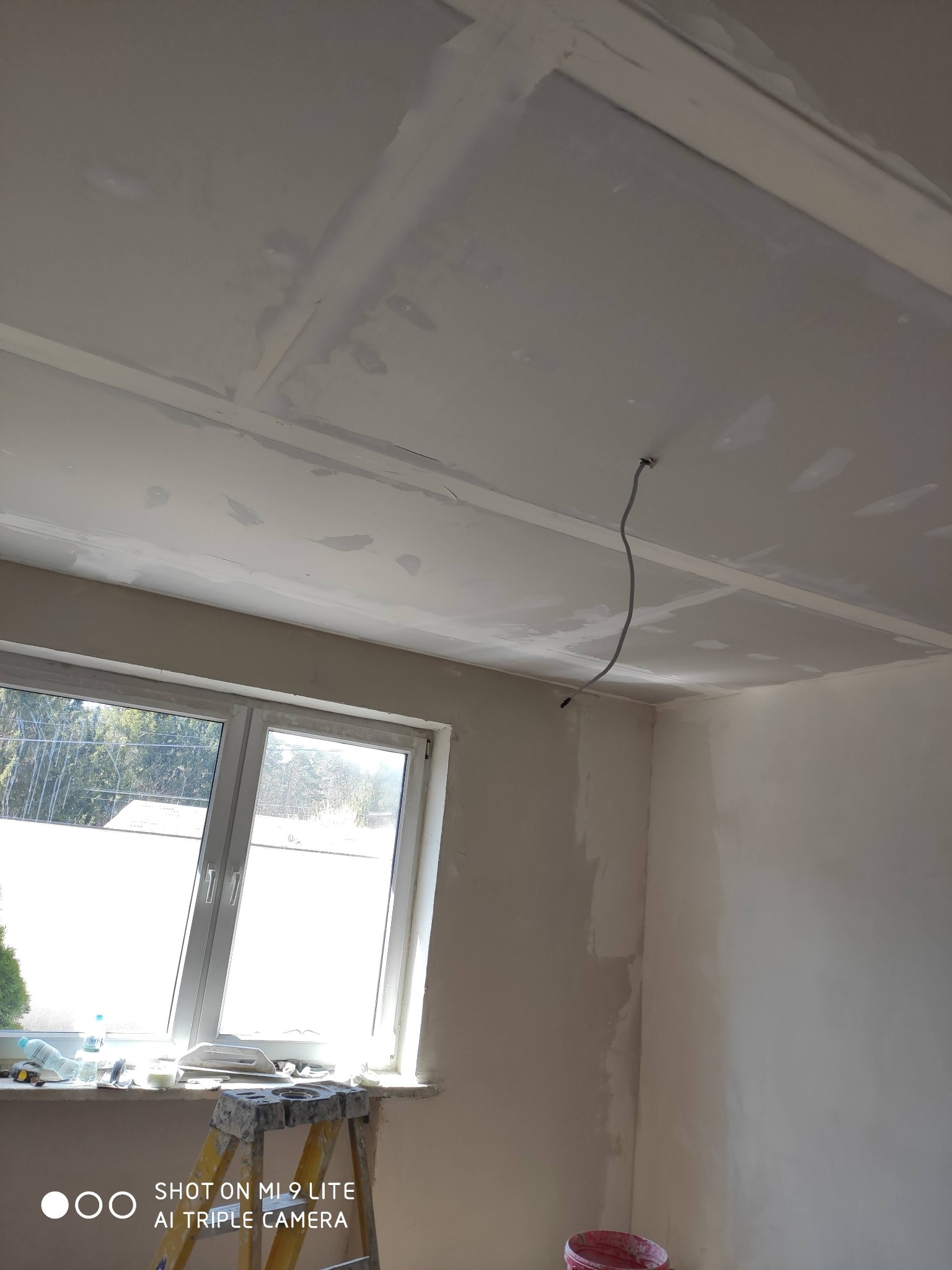 Pokój w trakcie prac wykończeniowych: ściany i sufit pokryte warstwą gładzi, widoczne łączenia płyt gipsowo-kartonowych, przewód elektryczny zwisający z sufitu, drabina przy oknie z narzędziami...