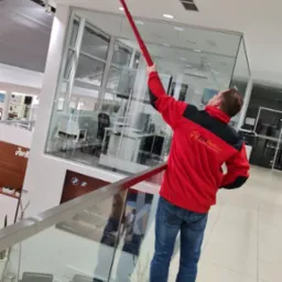 Mężczyzna w czerwonej bluzie z logo Fire Perfect czyści czujnik dymu na suficie w nowoczesnym biurze za pomocą specjalistycznego narzędzia z długim uchwytem.